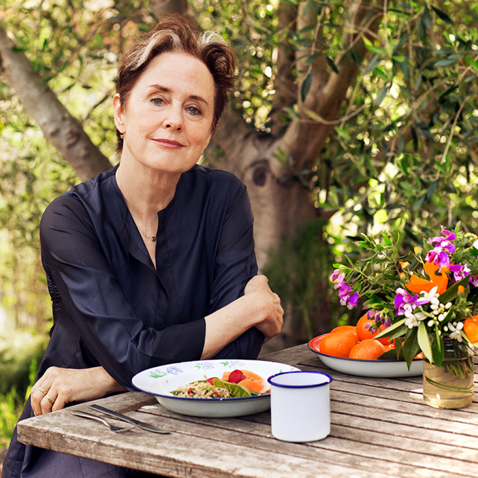 Photo of the real Alice Waters. She is sitting at a picnic table in front of a tree with a plate of fresh food. She is dressed in a black button-up shirt and sitting with her arms crossed.