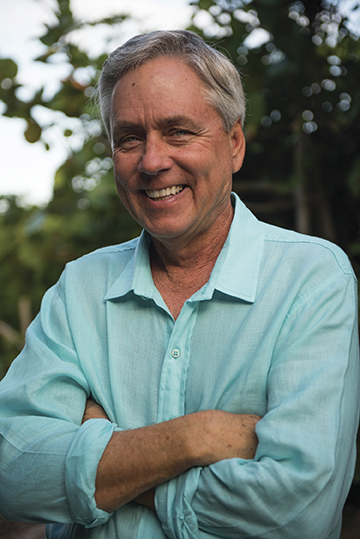 Portrait photo of author Carl Hiaasen, standing outside in an aqua button-up shirt with his arms crossed, smiling.