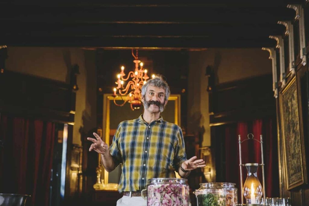 Photo of the real Sandor Katz. He is dressed in a yelllow and blue plaid shirt and is standing, gesturing at ingredients for making sauerkraut that are on a table in front of him.