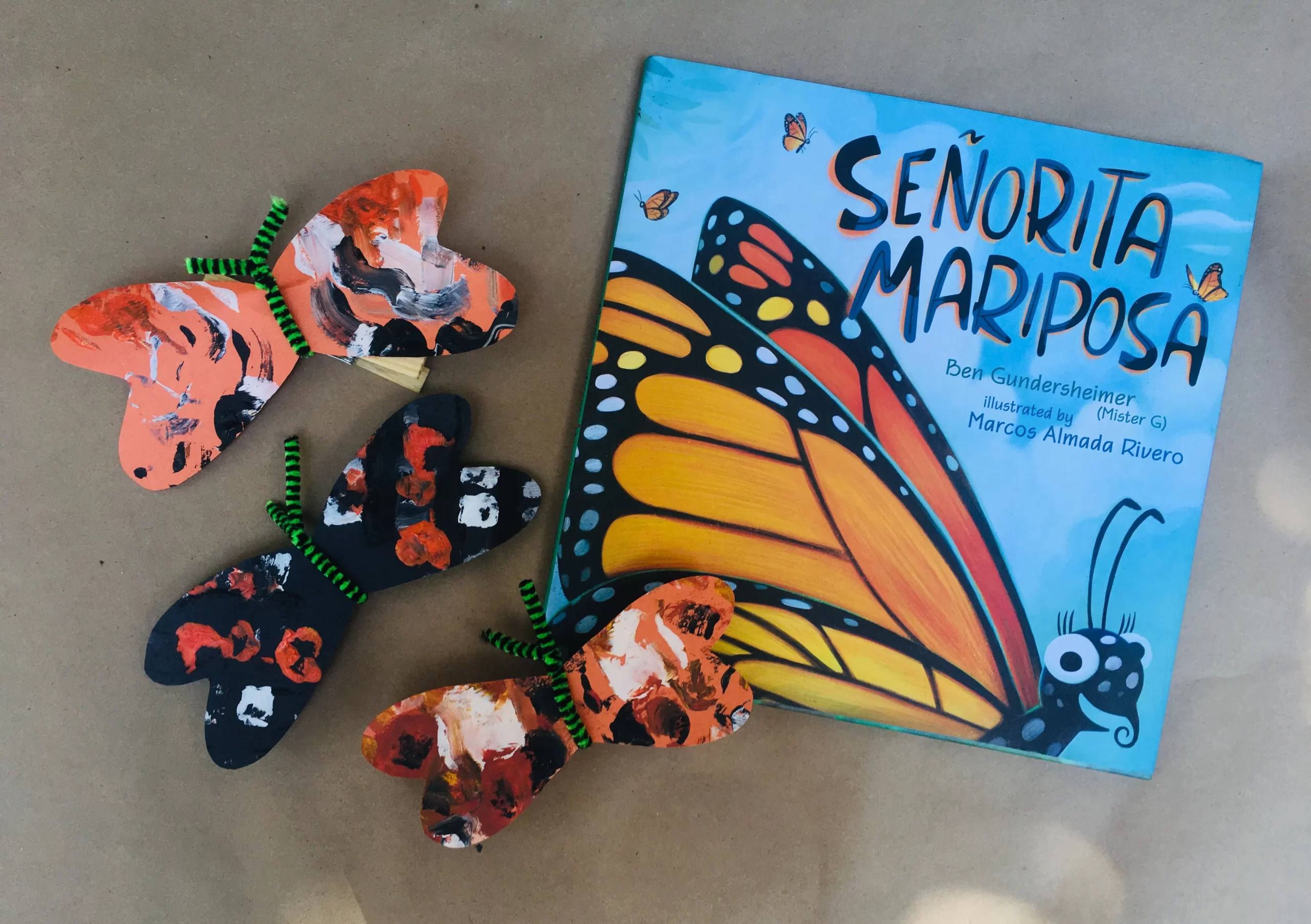 Cover of the book Señorita Mariposa, featuring a personified Monarch butterfly on a blue sky background. The book is surrounded by three examples of a butterfly craft.