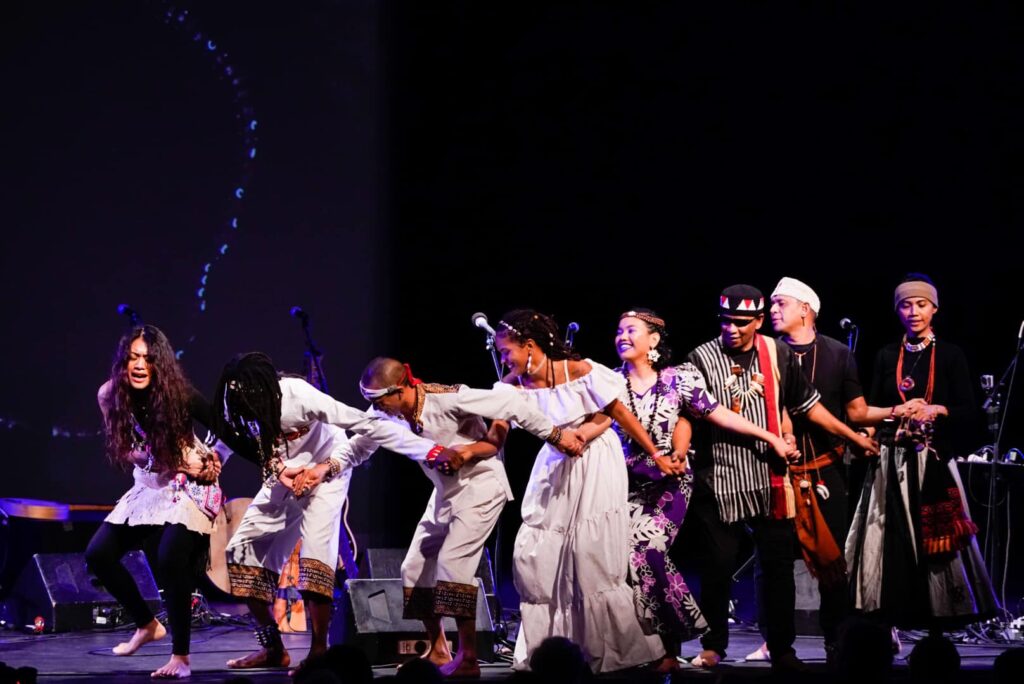 Photo of Small Island Big Song performing. There are 8 performers on stage who are positioned in a line with interlinking arms. They are bending over at their waists and dancing together. Some have expressions of joy, others of pain, and others are looking at the ground.
